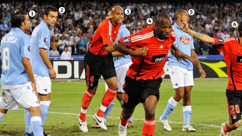 Cầu thủ Benfica với áo đỏ đang chống đỡ cầu thủ Napoli với áo xanh trên sân cỏ